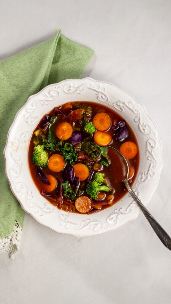 an overhead photo of a bowl of soup containing carrots, broccoli kale in a tomato broth. The bowl sits on a green napkin. A silver spoon sits inside the bowl.