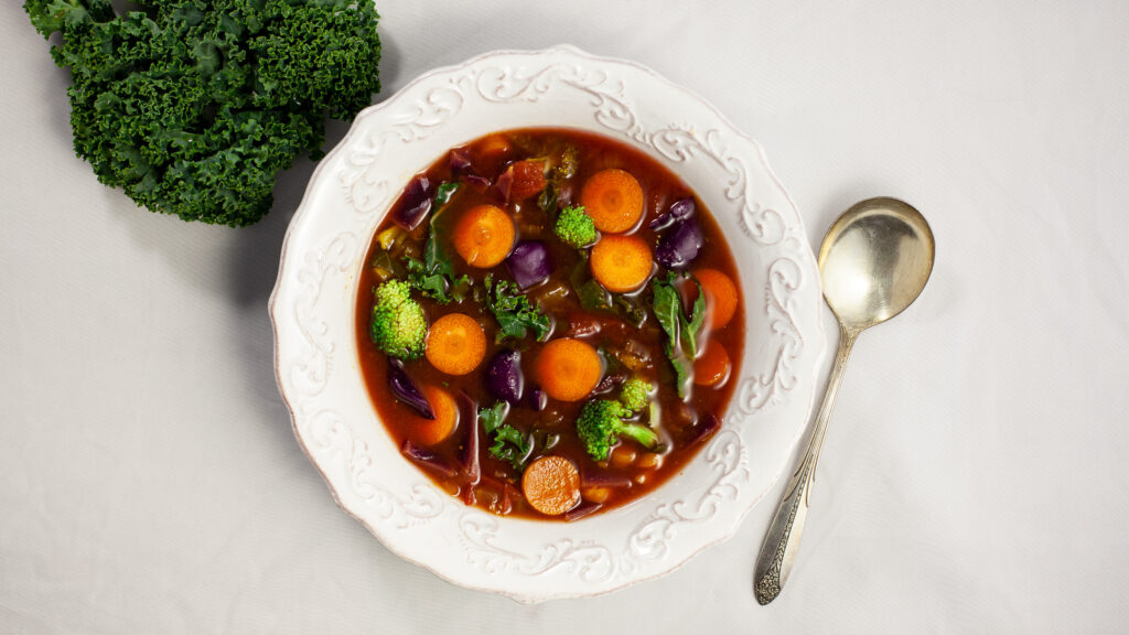 an overhead photo of a bowl of soup containing carrots, broccoli kale in a tomato broth. The bowl sits next to a kale leaf. A silver spoon sits beside the bowl.