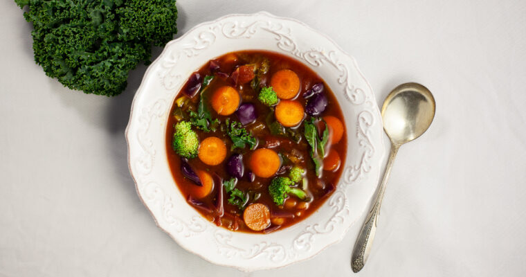 an overhead photo of a bowl of soup containing carrots, broccoli kale in a tomato broth. The bowl sits next to a kale leaf. A silver spoon sits beside the bowl.
