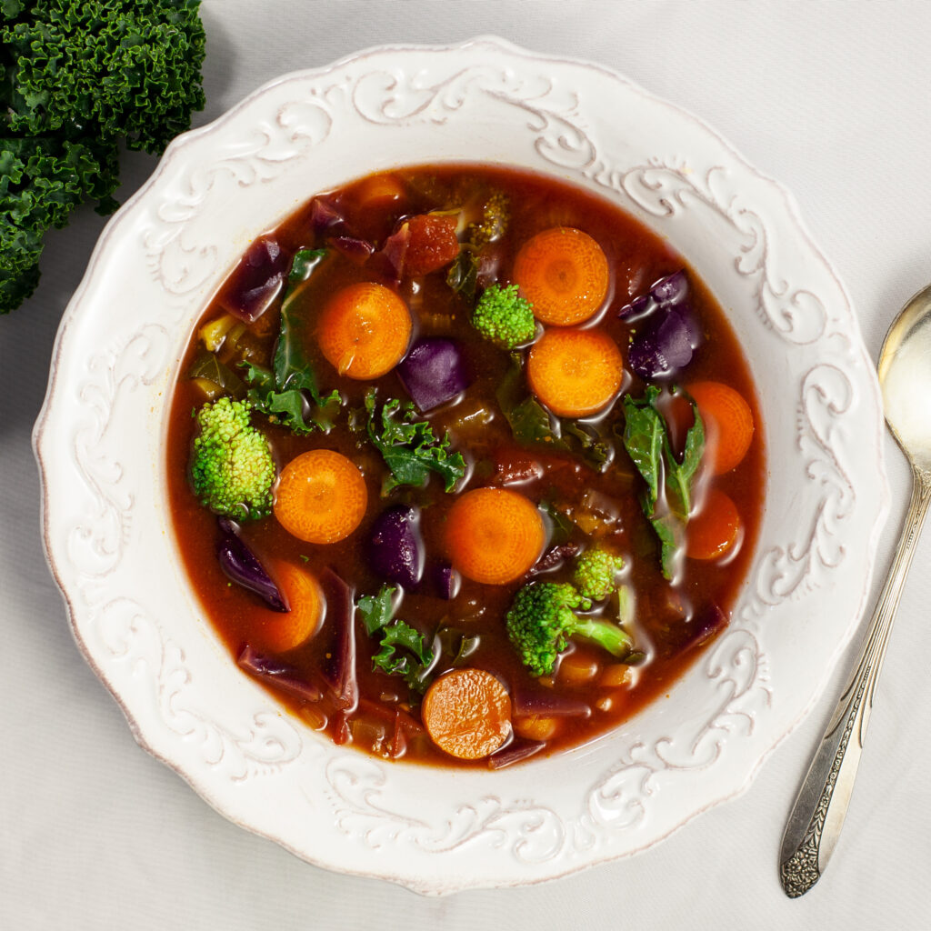 an overhead photo of a bowl of soup containing carrots, broccoli kale in a tomato broth. The bowl sits next to a kale leaf. A silver spoon sits beside the bowl.