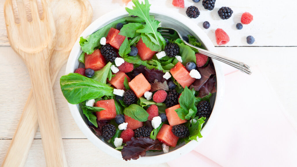 In a white bowl, a green salad has cubes of watermelon, fresh blackberries, fresh blueberries, chunks of feta and a balsamic dressing. Next to the bowl is the fruit.