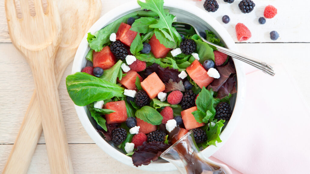 In a white bowl, a green salad has cubes of watermelon, fresh blackberries, fresh blueberries, chunks of feta and a balsamic dressing. Next to the bowl is the fruit.,