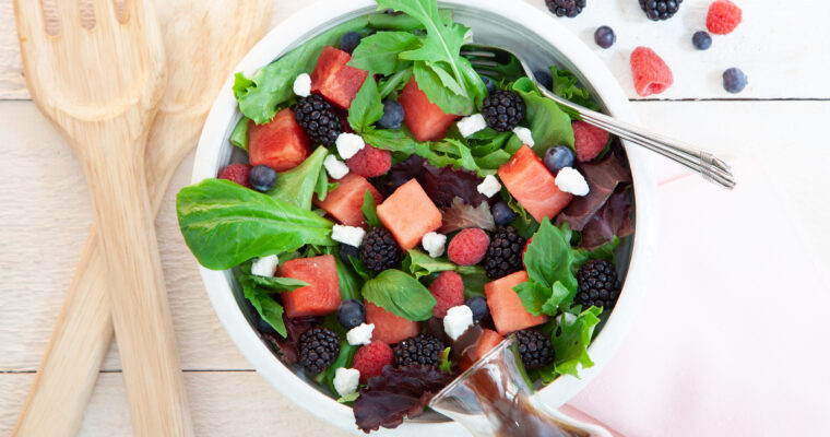 In a white bowl, a green salad has cubes of watermelon, fresh blackberries, fresh blueberries, chunks of feta and a balsamic dressing. Next to the bowl is the fruit.,