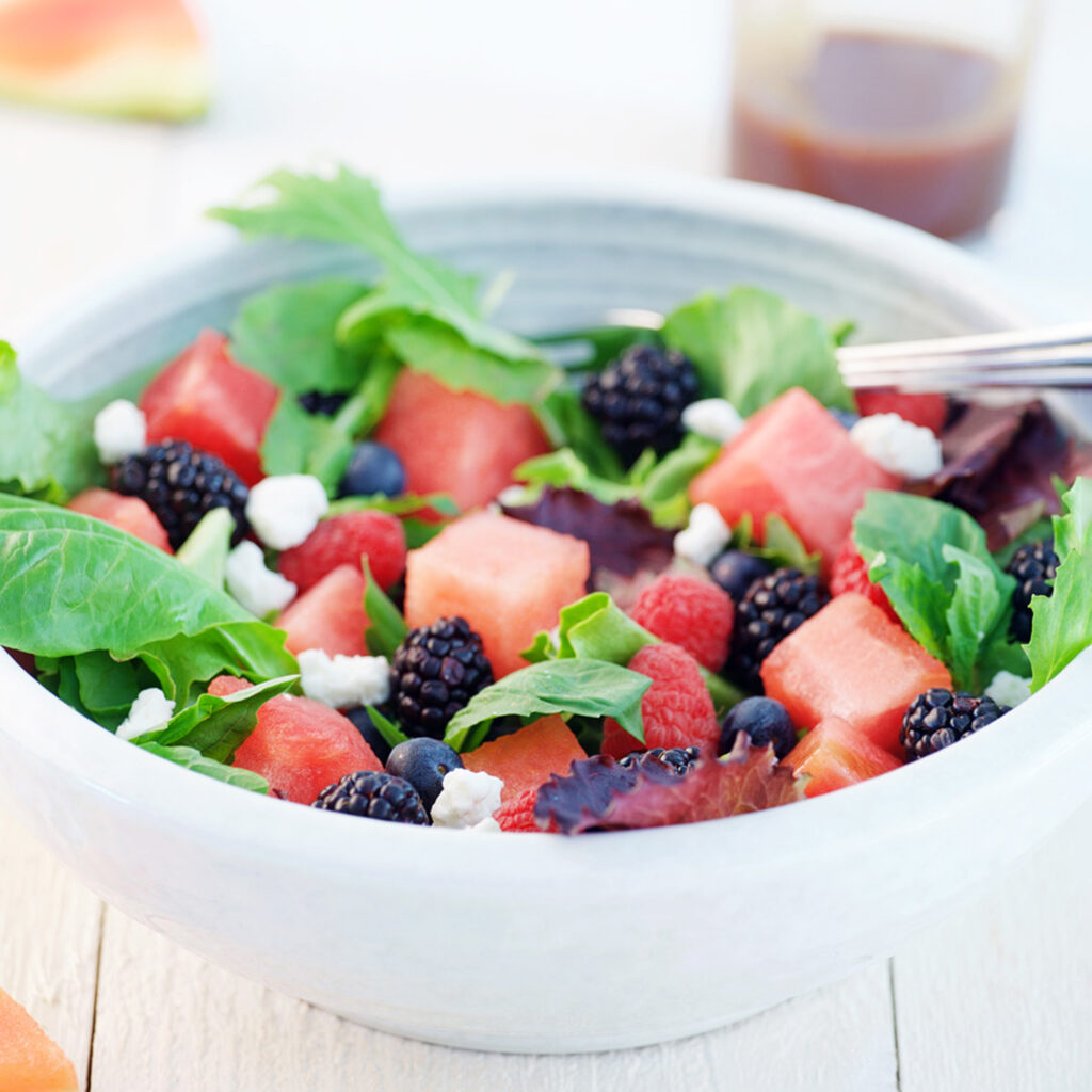 In a white bowl, a green salad has cubes of watermelon, fresh blackberries, fresh blueberries, chunks of feta and a balsamic dressing. Next to the bowl is the fruit.