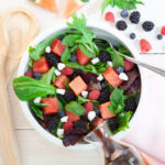 In a white bowl, a green salad has cubes of watermelon, fresh blackberries, fresh blueberries, chunks of feta and a balsamic dressing. Next to the bowl is the fruit.