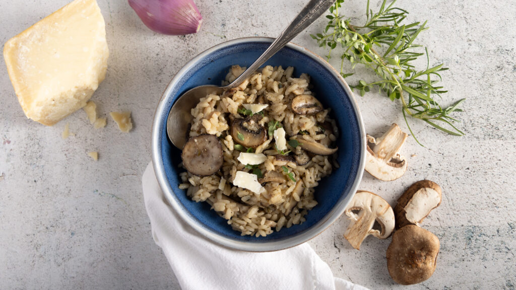 An overhead view of a bowl of wild mushroom risotto, with mushrooms, parmesan cheese, a shallot, fresh thyme and rosemary, and a white dinner napkin.