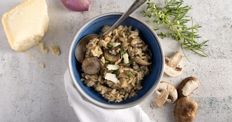 Photo alt: An overheads view of a bowl of wild mushroom risotto, with mushrooms, parmesan cheese, a shallot, fresh thyme and rosemary, and a white dinner napkin.