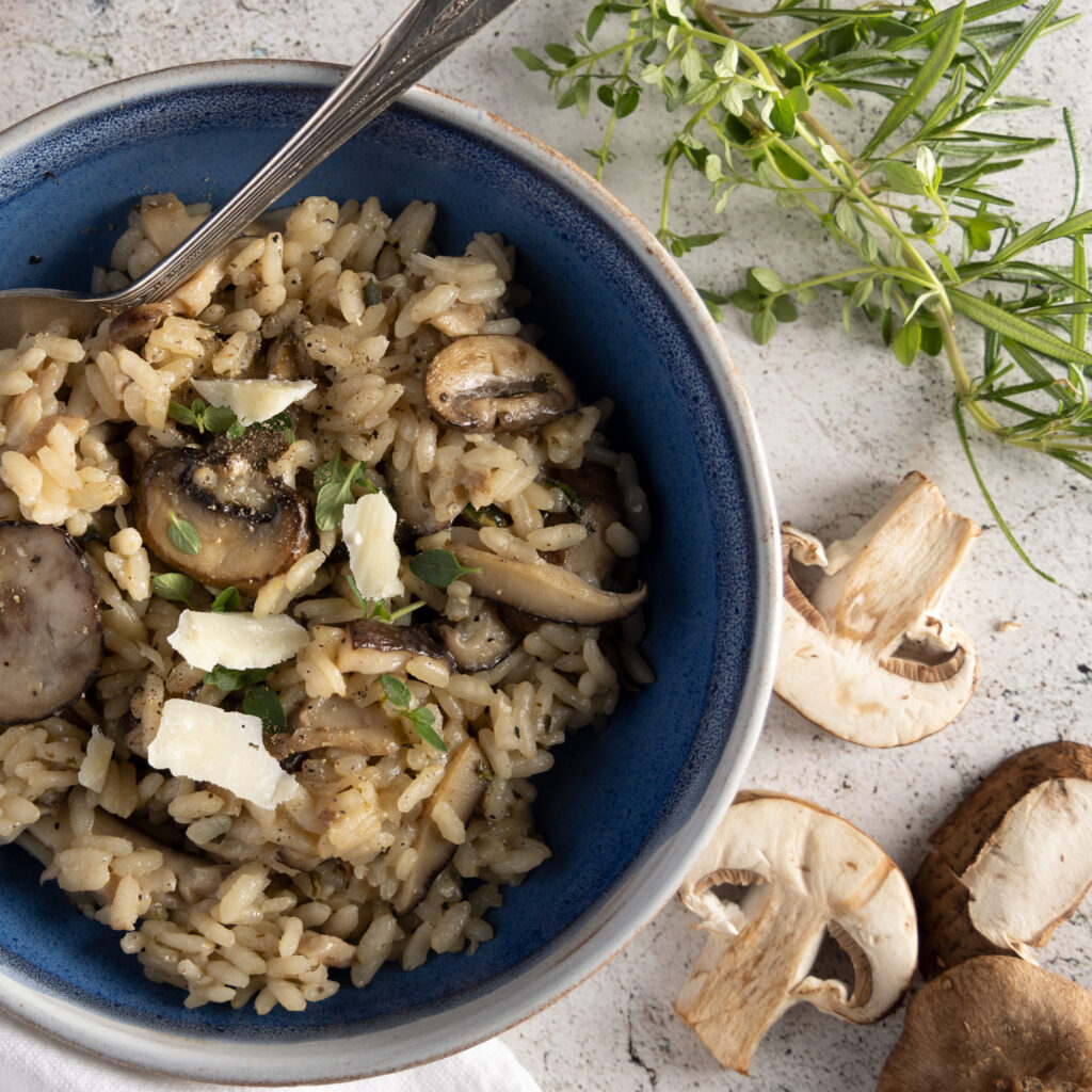 An overhead view of a bowl of wild mushroom risotto, with mushrooms, parmesan cheese, a shallot, fresh thyme and rosemary.