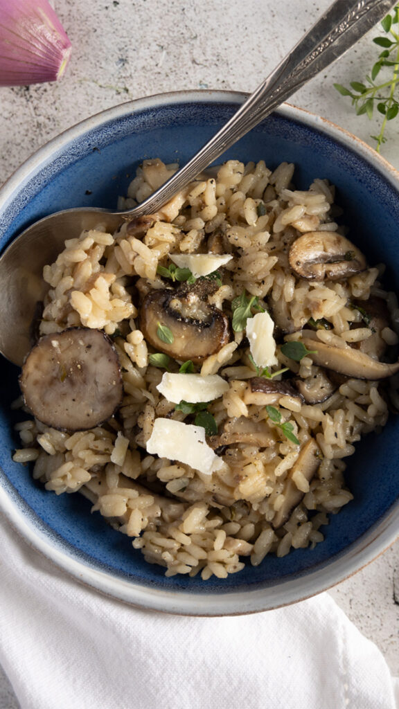 An overhead view of a bowl of wild mushroom risotto, with a shallot, fresh thyme and rosemary, and a white dinner napkin.