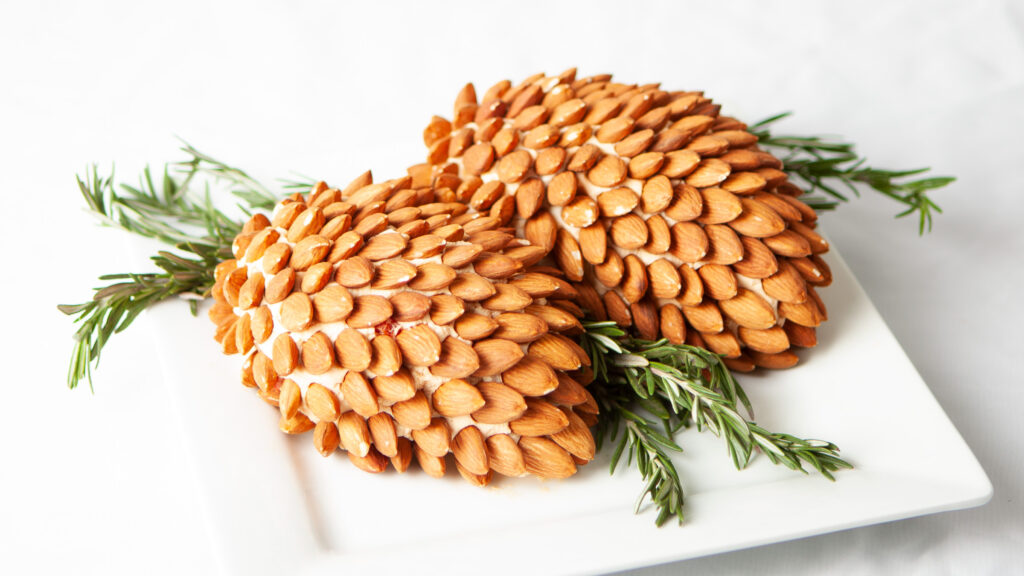 A photo of two pinecone-shaped cheddar spreads, covered with almonds, garnished with rosemary sprigs.