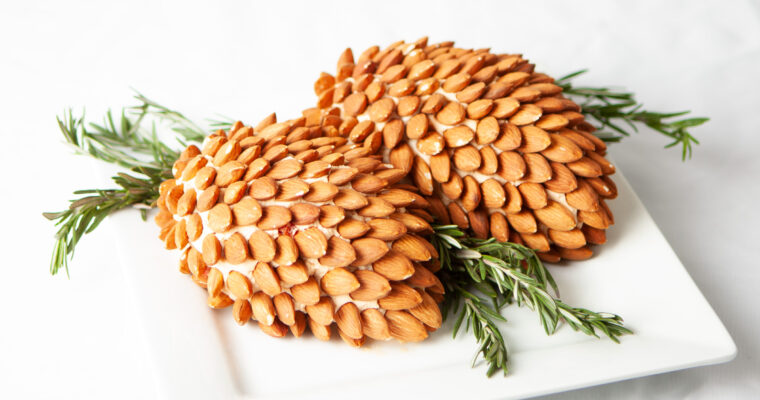 A photo of two pinecone-shaped cheddar spreads, covered with almonds, garnished with rosemary sprigs.