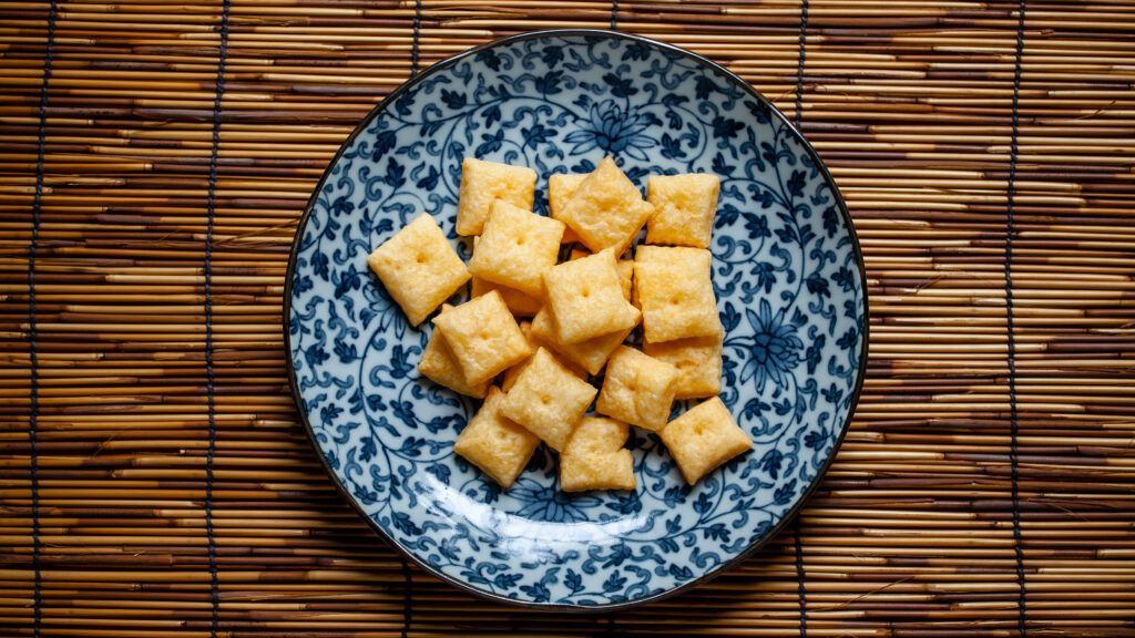 An overhead photo of a blue and white plate, with many tiny square cheese crackers. The plate is on a bamboo placemat.