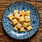 An overhead photo of a blue and white plate, with many tiny square cheese crackers. The plate is on a bamboo placemat.