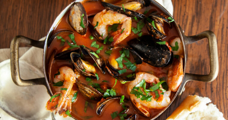 An overhead photo of a copper pot containing cioppino seafood stew, you can see mussels, prawns clams, in a tomato broth, garnished with fresh Italian parsley. The pot sits on a cream coloured napkin. A chunk of crusty bread sits beside the pot. The scene is on a wooden table.