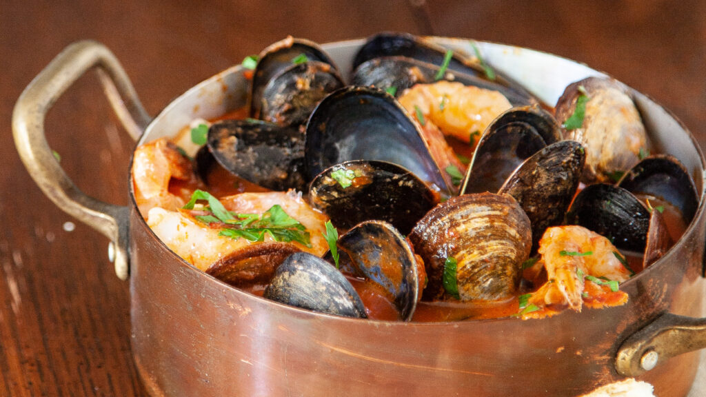 A three-quarter view photo of a copper pot containing cioppino seafood stew, you can see mussels, prawns clams, in a tomato broth, garnished with fresh Italian parsley. The scene is on a wooden table.