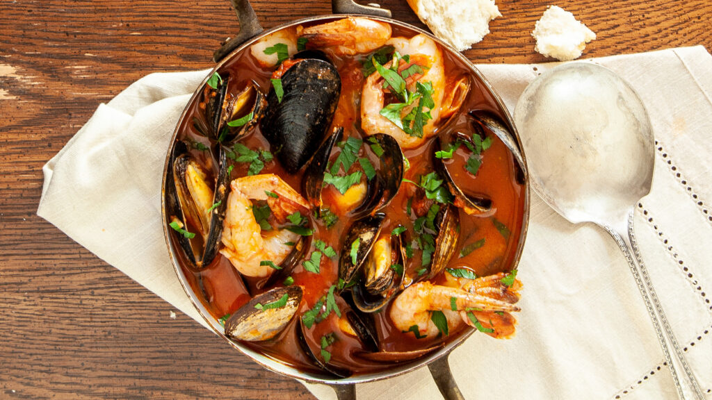 An overhead photo of a copper pot containing cioppino seafood stew, you can see mussels, prawns clams, in a tomato broth, garnished with fresh Italian parsley. The pot sits on a cream coloured napkin. A chunk of crusty bread sits beside the pot. The scene is on a wooden table.