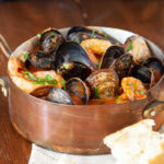 An overhead photo of a copper pot containing cioppino seafood stew, you can see mussels, prawns clams, in a tomato broth, garnished with fresh Italian parsley. The pot sits on a cream coloured napkin. A chunk of crusty bread sits beside the pot. The scene is on a wooden table.