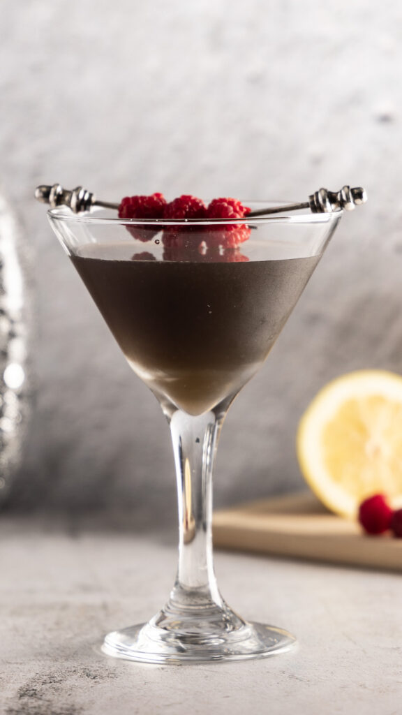 A photo of a dark-coloured martini in a martini glass, with lemon and raspberries in the background, a silver sequined pumpkin, and a crow decoration.