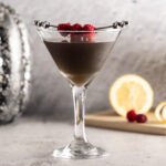 A photo of a dark-coloured martini in a martini glass, with lemon and raspberries in the background, a silver sequined pumpkin, and a crow decoration.