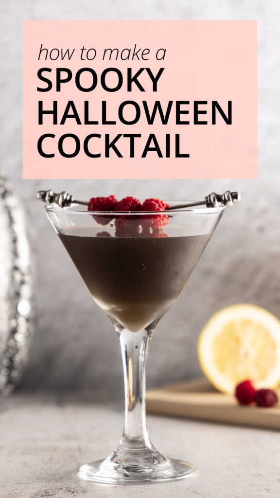 A photo of a dark-coloured martini in a martini glass, with lemon and raspberries in the background, a silver sequined pumpkin, and a crow decoration.