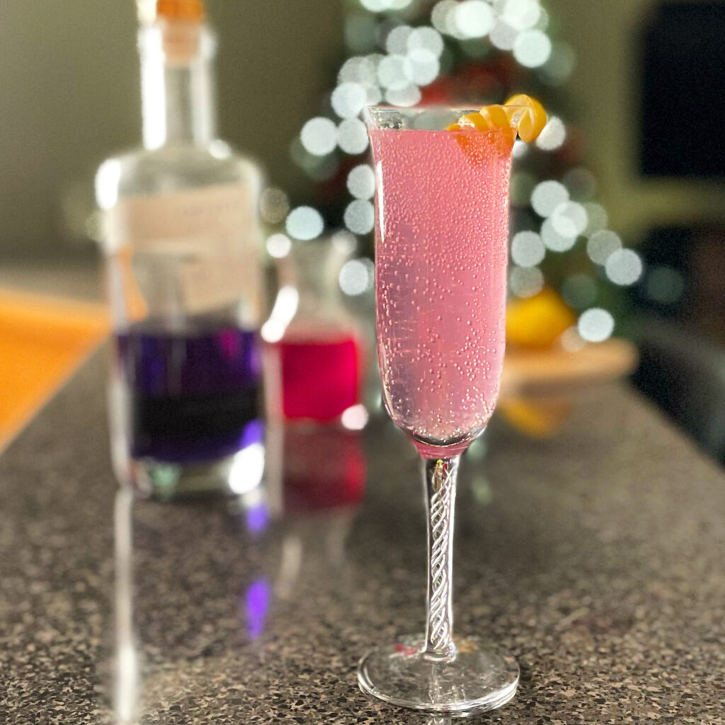 A photo of everything needed to put into a Pink 75 cocktail; champagne flute glass, cocktail shakerm measuring cups, lemon, Empress 1908 gin in the bottle, and a vial of red berry syurup.