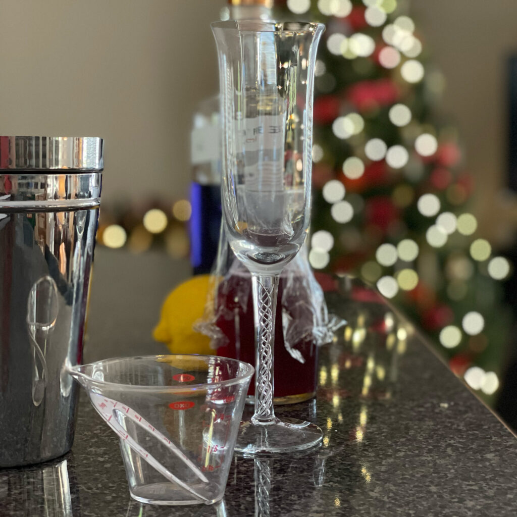 A photo of everything needed to put into a Pink 75 cocktail; champagne flute glass, cocktail shakerm measuring cups, lemon, Empress 1908 gin in the bottle, and a vial of red berry syurup.