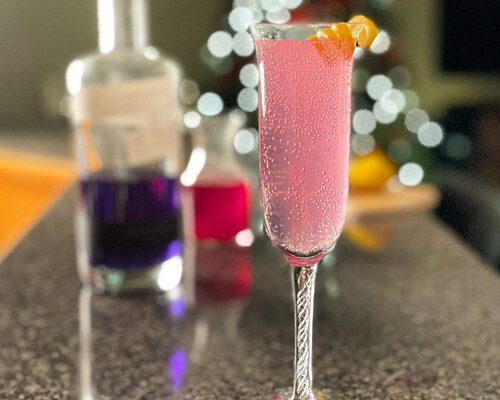 A photo of everything needed to put into a Pink 75 cocktail; champagne flute glass, cocktail shaker, measuring cups, lemon, Empress 1908 gin in the bottle, and a vial of red berry syrup.