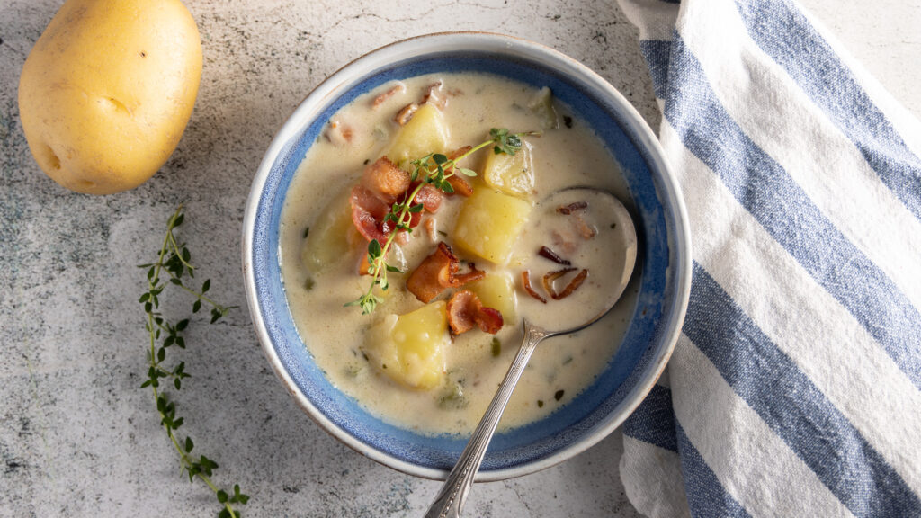 An overhead photo of a bowl of creamy potato and bacon soup, with a soup spoon, blue and white napkin, yellow potatoes, and bacon. A spring of thyme is the garnish on the soup.