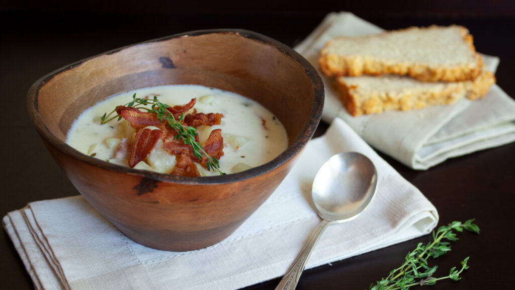 A photo of a wooden bowl filled with creamy potato and bacon soup, with a soup spoon, white napkin, garnished with pieces of bacon and fresh thyme sprigs. Two slices of crusty bread are in the photo.