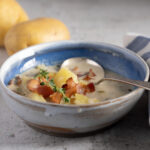 A photo of a bowl of creamy potato and bacon soup, with a soup spoon, blue and white napkin, yellow potatoes, and bacon. A spring of thyme is the garnish on the soup.