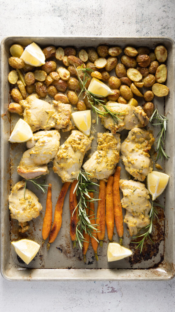 An overhead photo of a sheet pan with chicken thighs, small whole carrots, small new potatoes, and sprigs of fresh rosemary.