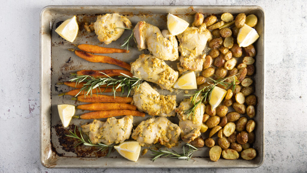 An overhead photo of a sheet pan with chicken thighs, small whole carrots, small new potatoes, and sprigs of fresh rosemary.