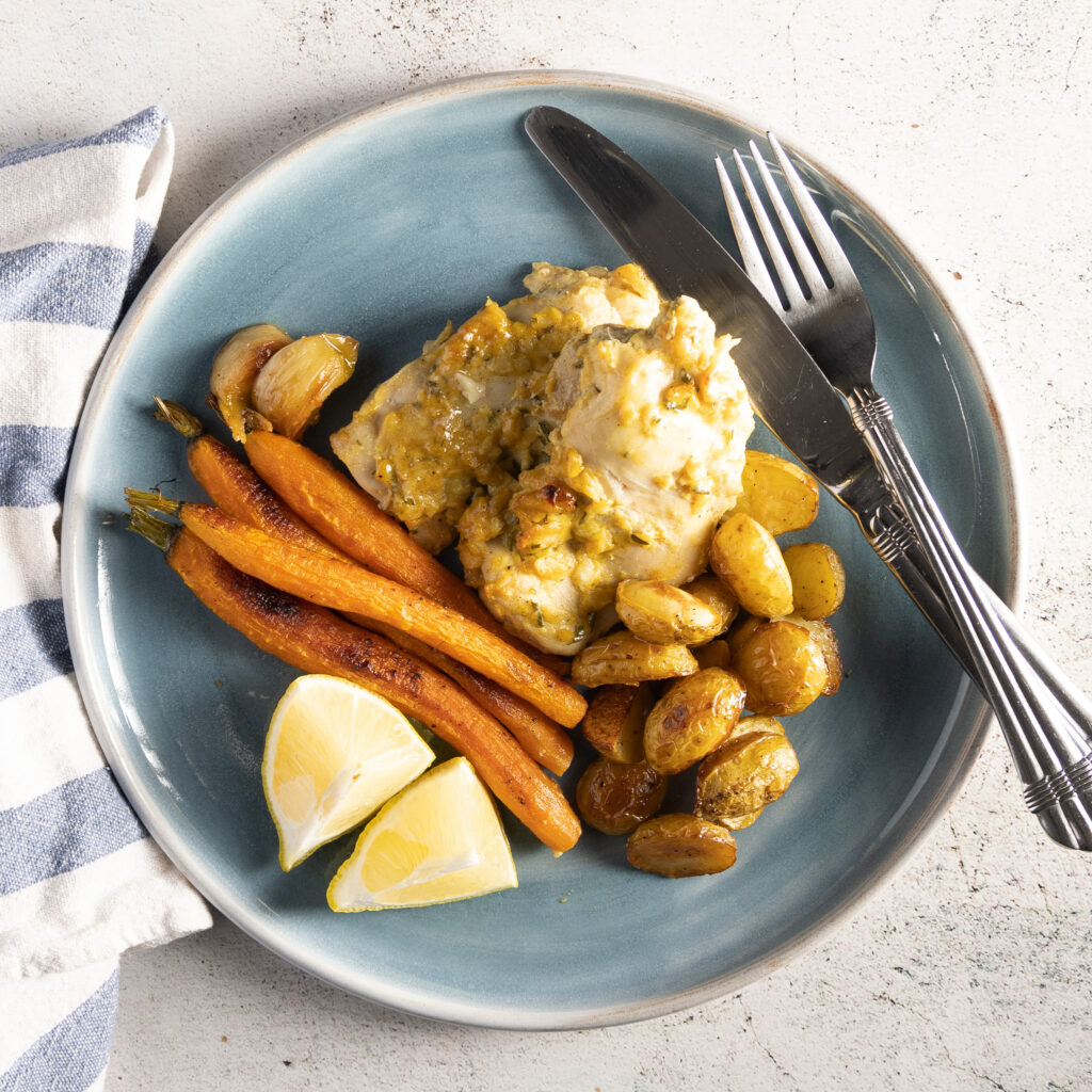 An overhead photo of a blue plate, with chicken thighs, small whole carrots, small new potatoes, and sprigs of fresh rosemary. A blue and white napkin sits next to the plate, and a silver knife and fork rest next to the food on the plate.