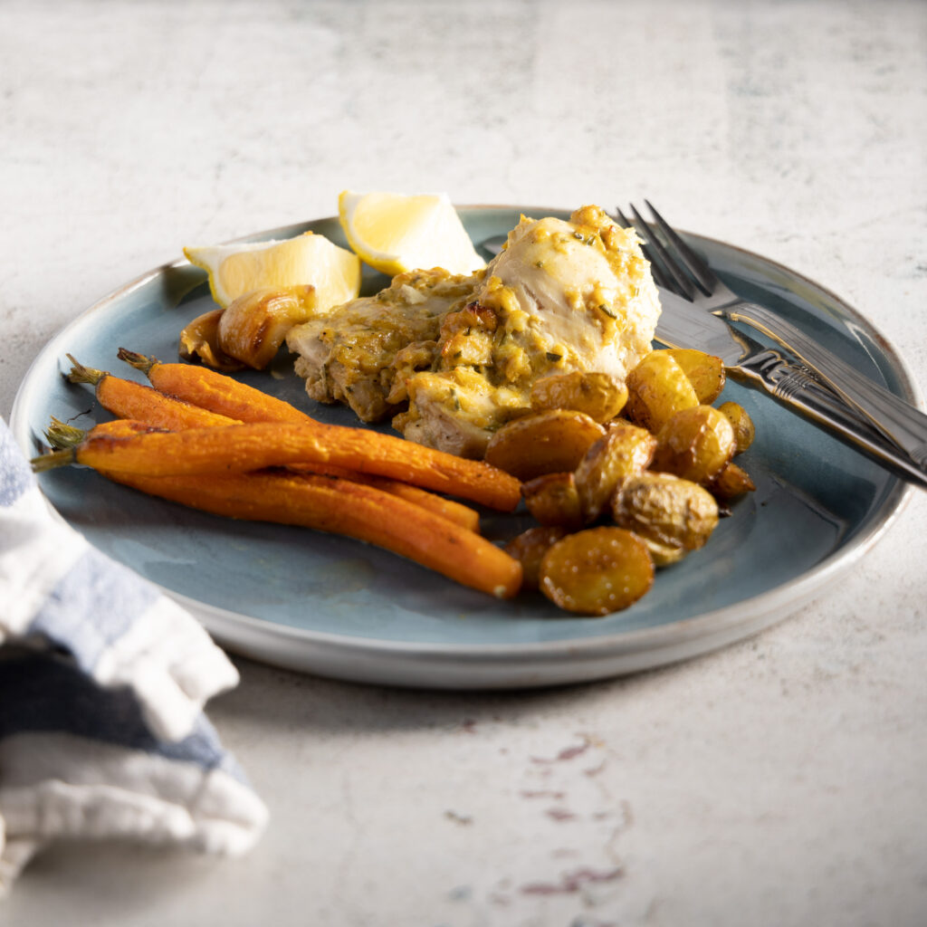 An three-quarter view photo of a blue plate, with chicken thighs, small whole carrots, small new potatoes, and sprigs of fresh rosemary. A blue and white napkin sits next to the plate, and a silver knife and fork rest next to the food on the plate.