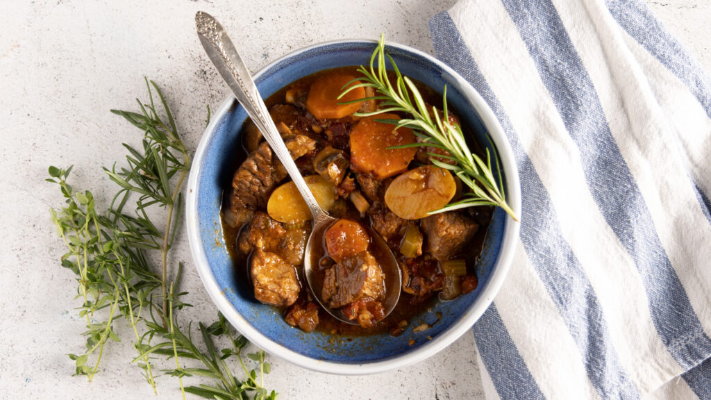 An overhead view of a bowl of beef stew, showing carrots, potatoes, and beef. A spoon rests in the bowl. Next to the bowl are fresh thyme and rosemary sprigs, and a blue and white striped towel.