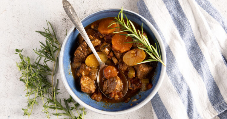 An overhead view of a bowl of beef stew, showing carrots, potatoes, and beef. A spoon rests in the bowl. Next to the bowl are fresh thyme and rosemary sprigs, and a blue and white striped towel.
