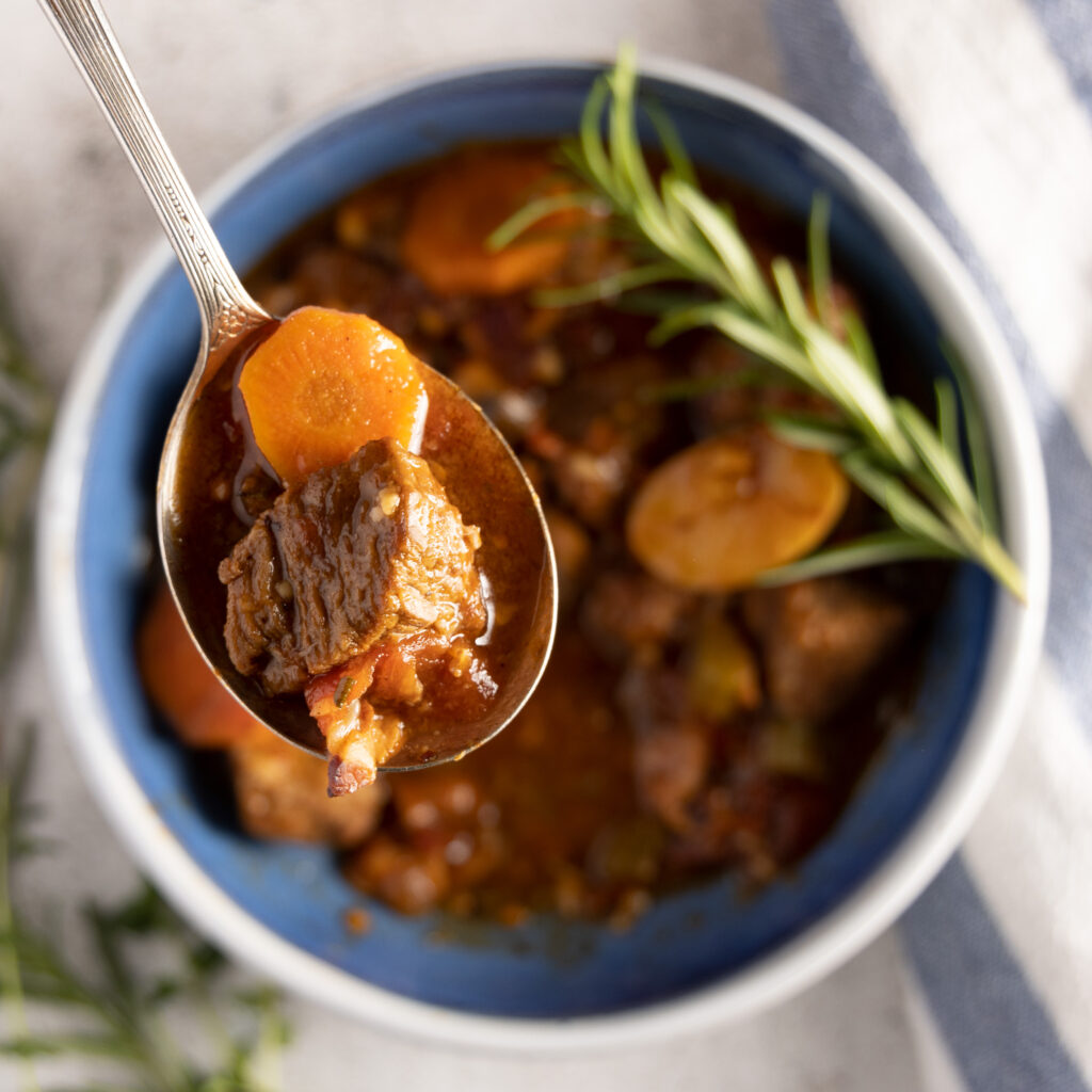 An overhead view of a bowl of beef stew, showing carrots, potatoes, and beef. A spoon sits above the bowl, containing a bite of beef and carrot. Next to the bowl are fresh thyme and rosemary sprigs, and a blue and white striped towel.