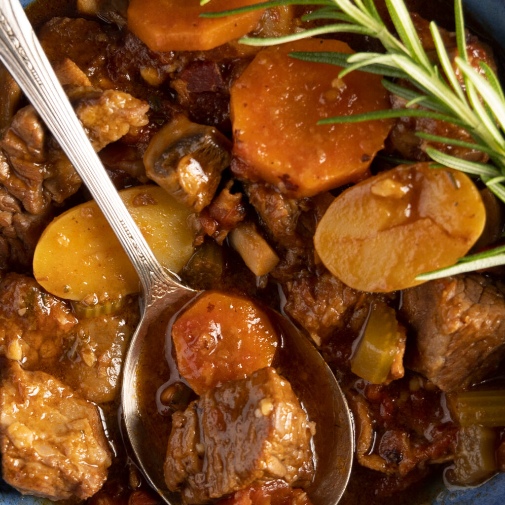 A zoomed in view of a spoon holding a bite of beef stew, showing a piece of steak, carrot, and showing a sprig of rosemary.