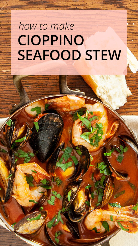 An overhead photo of a copper pot containing cioppino seafood stew, you can see mussels, prawns clams, in a tomato broth, garnished with fresh Italian parsley. The pot sits on a cream coloured napkin. A chunk of crusty bread sits beside the pot. The scene is on a wooden table.