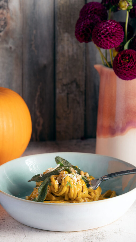 A bowl of pumpkin pasta topped with crispy sage leaves and parmesan cheese sits on a table, next to two whole pumpkins, and a pink vessel containing purple dahlia flowers. 