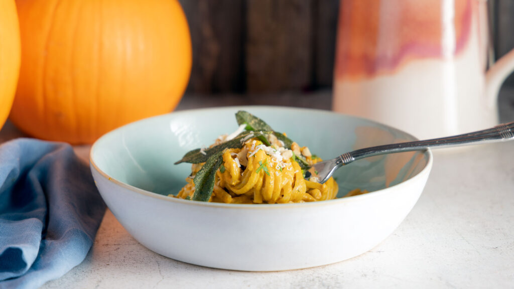 A bowl of pumpkin pasta topped with crispy sage leaves and parmesan cheese sits on a table, next to two whole pumpkins, a pink vessel, a blue napkin. 