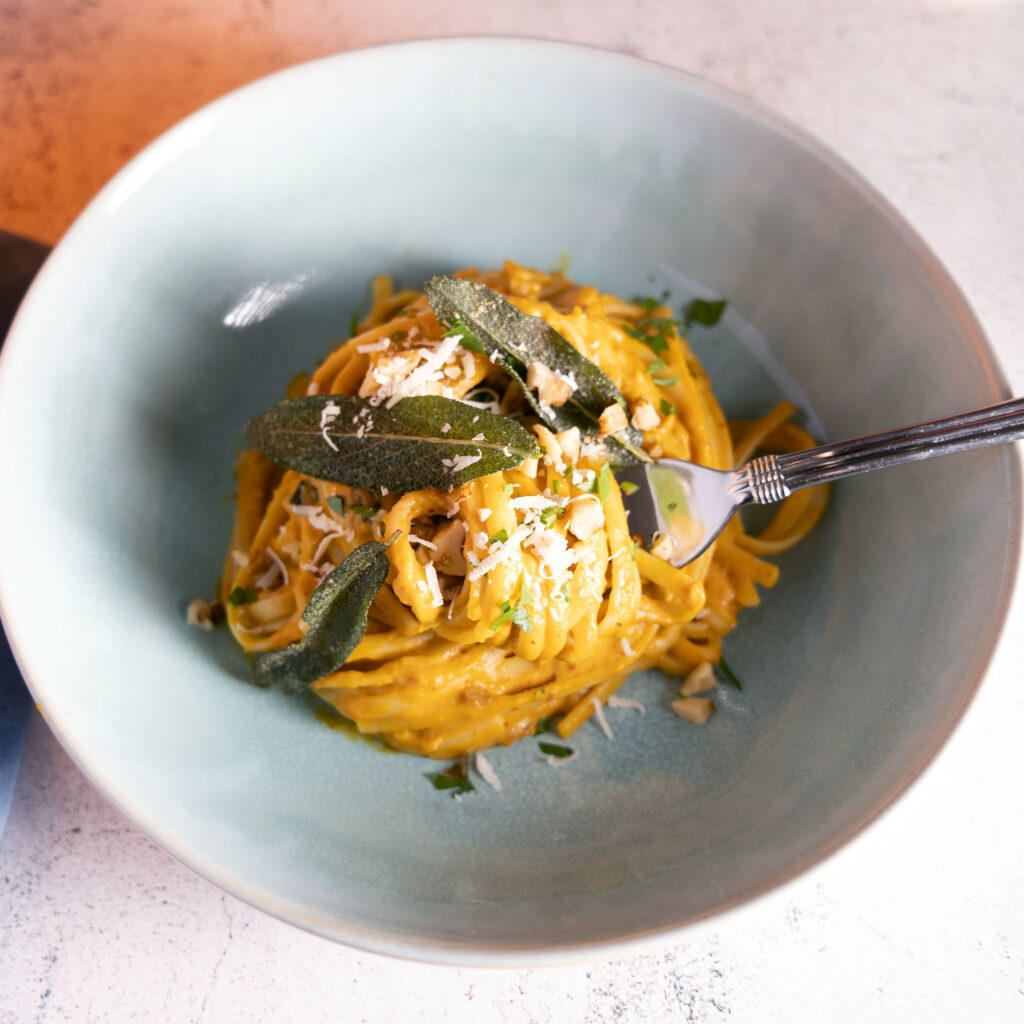 A bowl of pumpkin pasta topped with crispy sage leaves and parmesan cheese sits on a table.