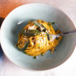 A bowl of pumpkin pasta topped with crispy sage leaves and parmesan cheese sits on a table.