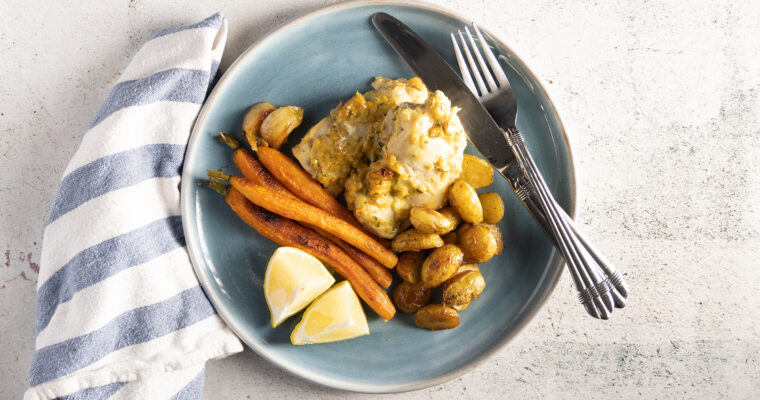 An overhead photo of a blue plate, with chicken thighs, small whole carrots, small new potatoes, and sprigs of fresh rosemary. A blue and white napkin sits next to the plate, and a silver knife and fork rest next to the food on the plate.