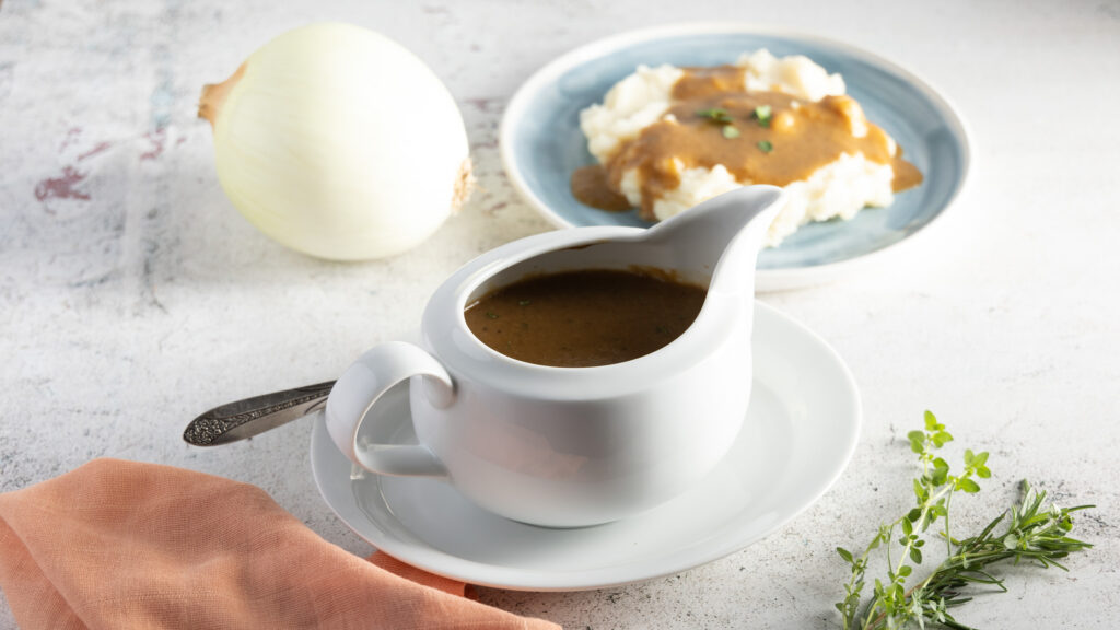 A photo of a white gravy boat and plate, with a silver coloured spoon. Inside the gravy boat is gravy. Behind the gravy boat is a blue plate with mashed potatoes, and gravy on top, with thyme leaves. A raw onion sits to the side.