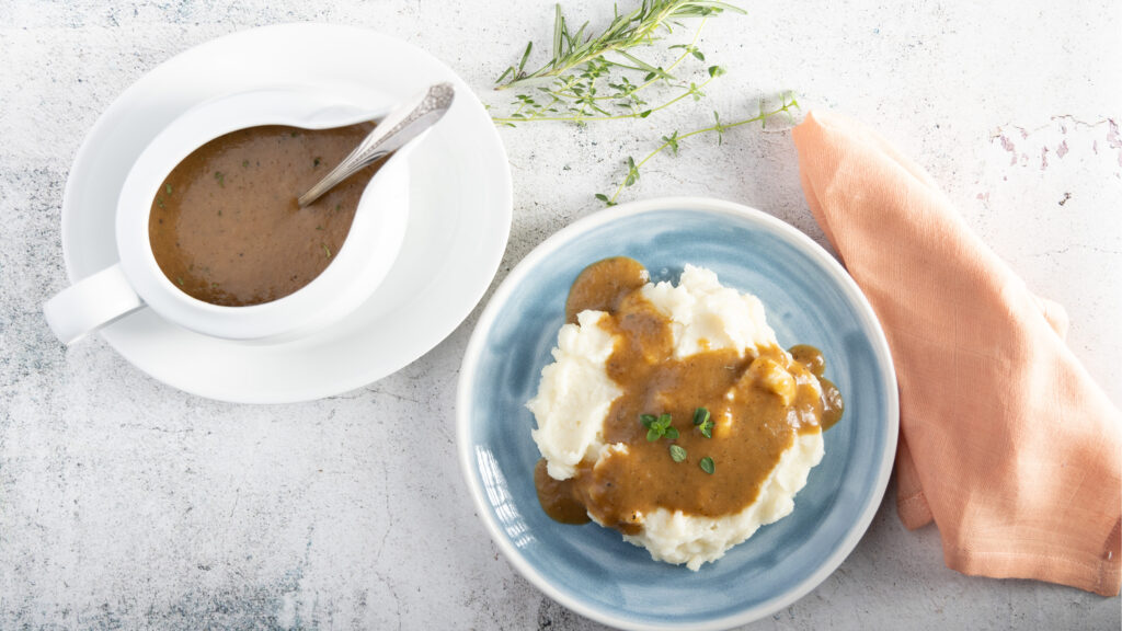 An overhead photo of a white gravy boat and plate, with a silver coloured spoon. Inside the gravy boat is gravy. Beside the gravy boat is a blue plate with mashed potatoes, and gravy on top, with thyme leaves and rosemary sprigs next to the plate. 