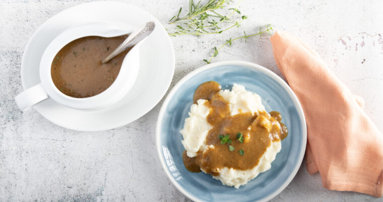 An overhead photo of a white gravy boat and plate, with a silver coloured spoon. Inside the gravy boat is gravy. Beside the gravy boat is a blue plate with mashed potatoes, and gravy on top, with thyme leaves and rosemary sprigs next to the plate.