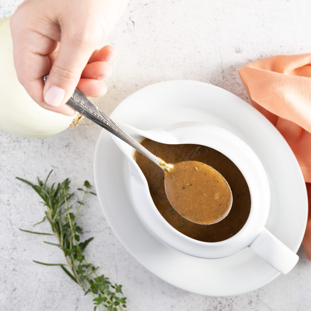 An overhead photo of a white gravy boat and plate, with a silver coloured spoon. Inside the gravy boat is gravy. Beside the gravy boat are thyme leaves and rosemary sprigs next to the plate. 