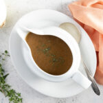 An overhead photo of a white gravy boat and plate, with a silver coloured spoon. Inside the gravy boat is gravy. Beside the gravy boat are thyme leaves and rosemary sprigs next to the plate.