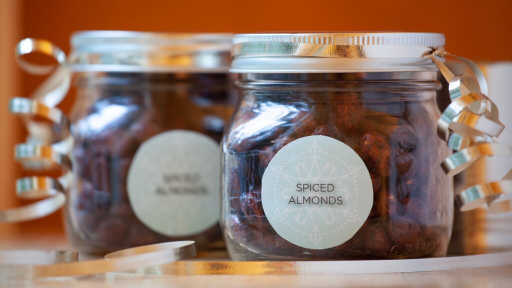 A pair of decorative jars contain holiday spiced nuts. The jars are decorated with silver ribbon and custom-labels that read, “Spiced Almonds”.